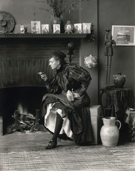 Frances Benjamin Johnston's Self-Portrait (as "New Woman"), a full-length self-portrait of her seated in front of fireplace, facing left, holding cigarette in one hand and a beer stein in the other, in her Washington, D.C. studio